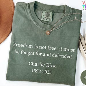 May include: A moss green t-shirt with the text "Freedom is not free; it must be fought for and defended" and "Charlie Kirk 1993-2025" printed in white. A gold necklace with a pendant rests on the shirt.