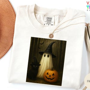 Puede incluir: Camiseta de color marfil con un gr&aacute;fico de Halloween. El gr&aacute;fico representa un fantasma con un sombrero de bruja, un gato negro con un sombrero de bruja y una calabaza tallada. La camiseta tiene una etiqueta Comfort Colors.