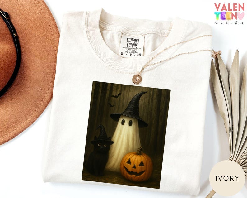 Puede incluir: Camiseta de color marfil con un gr&aacute;fico de Halloween. El gr&aacute;fico representa un fantasma con un sombrero de bruja, un gato negro con un sombrero de bruja y una calabaza tallada. La camiseta tiene una etiqueta Comfort Colors.