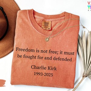 May include: A yam-colored Comfort Colors t-shirt with the quote "Freedom is not free; it must be fought for and defended" and the name Charlie Kirk, 1993-2025. A gold necklace and a brown hat are also visible.
