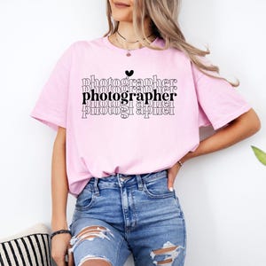 May include: A pink t-shirt with the word "photographer" repeated in a stacked design with a small heart above the first word. The shirt is being worn by a person with long brown hair.