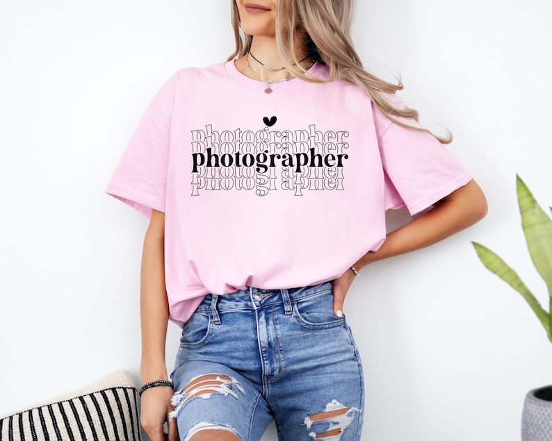 May include: A pink t-shirt with the word "photographer" repeated in a stacked design with a small heart above the first word. The shirt is being worn by a person with long brown hair.