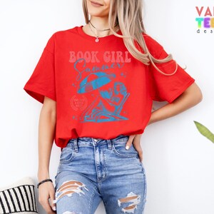 May include: A red t-shirt with a blue graphic that reads "BOOK GIRL Summer". The graphic depicts a person relaxing in a beach chair under an umbrella. The shirt is paired with blue jeans.