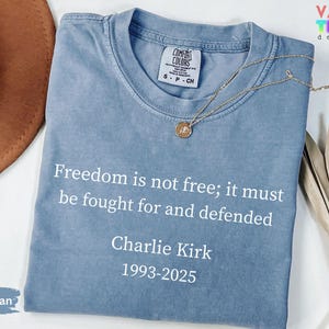 May include: A blue jean colored t-shirt with the text "Freedom is not free; it must be fought for and defended" and "Charlie Kirk 1993-2025" in white. A gold necklace with a pendant is on the shirt.