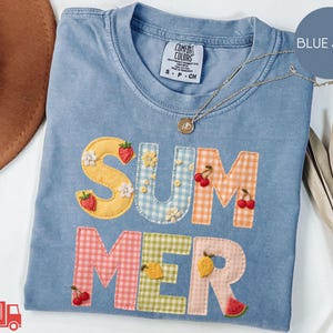 May include: Blue jean colored t-shirt with the word "SUMMER" appliqued on the front. The letters are in various patterns and colors, with fruit and flower embellishments. A gold necklace with a pendant is on the shirt.