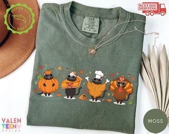 May include: A moss green crew neck t-shirt featuring a graphic of four black cats dressed in autumn-themed costumes, including a pumpkin, a slice of pie, a roasted turkey, and a turkey bird, surrounded by small fall leaves and acorns.