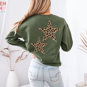 May include: An olive green sweatshirt featuring two leopard print stars on the back. The sweatshirt has long sleeves and a crew neck. The text "VALEN TEEN design" is visible in the upper left corner.
