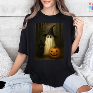 Puede incluir: Camiseta negra con un dise&ntilde;o de Halloween. El gr&aacute;fico muestra un fantasma con sombrero de bruja, un gato negro y una calabaza tallada. La camiseta tiene la palabra "Black" en la parte inferior.