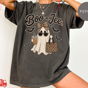 May include: A charcoal gray short-sleeve t-shirt featuring a graphic of a ghost wearing sunglasses and a leopard print bow, holding a coffee cup and a leopard print tote bag. The text Boo-Jee is printed above the ghost, surrounded by black bats.