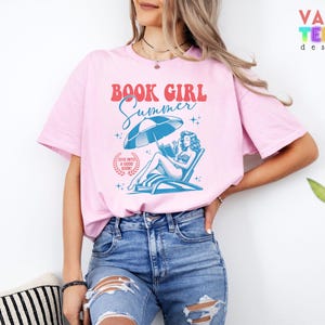May include: Pink t-shirt with the text "BOOK GIRL Summer" in red and blue. The graphic features a woman reading under an umbrella on a beach chair. The shirt also has the text "DIVE INTO A GOOD BOOK".
