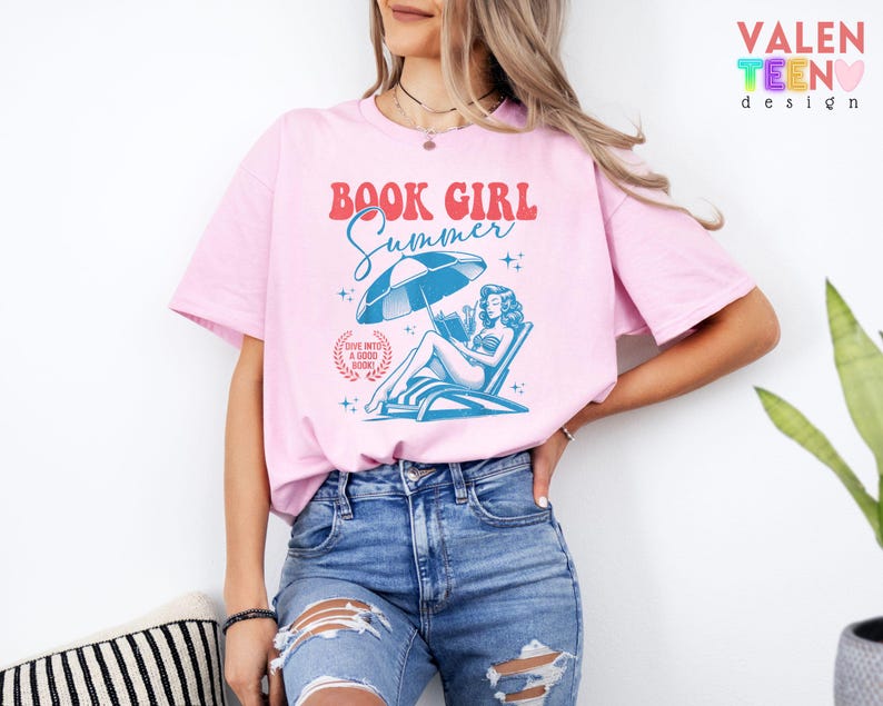 May include: Pink t-shirt with the text "BOOK GIRL Summer" in red and blue. The graphic features a woman reading under an umbrella on a beach chair. The shirt also has the text "DIVE INTO A GOOD BOOK".