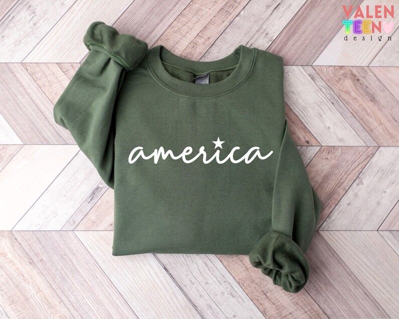 Puede incluir: Una sudadera verde oliva con la palabra "america" en escritura blanca, con una estrella encima de la "i". Las mangas est&aacute;n dobladas. La sudadera se muestra sobre una superficie de madera marr&oacute;n claro. La marca "VALENTEEN design" est&aacute; en la esquina superior derecha.