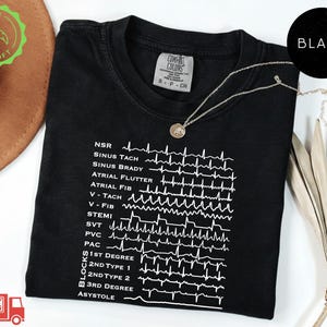 May include: A black crew neck t-shirt featuring a white graphic print of various cardiac rhythm strips and medical abbreviations including NSR, Sinus Tach, Atrial Flutter, V-Fib, STEMI, and Asystole, with a Comfort Colors brand label at the neck.