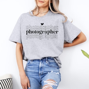 May include: A gray t-shirt with the word "photographer" repeated in a stacked pattern. The word "photographer" is in black and the rest of the text is in gray. A small black heart is above the word "photographer".