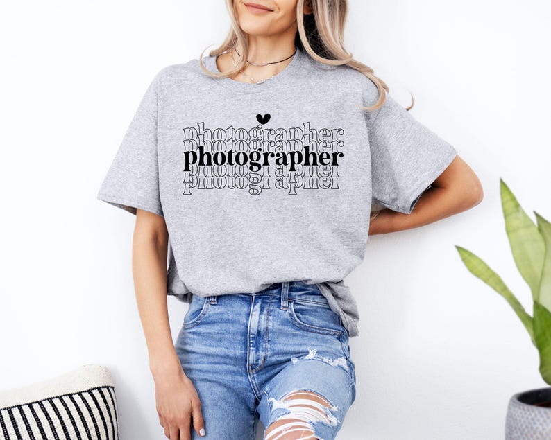May include: A gray t-shirt with the word "photographer" repeated in a stacked pattern. The word "photographer" is in black and the rest of the text is in gray. A small black heart is above the word "photographer".