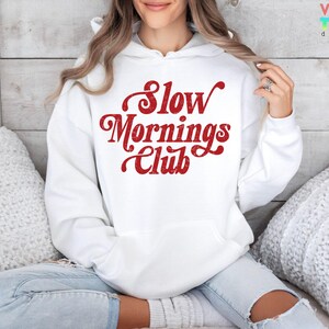 May include: White hoodie with a front pocket and the text "Slow Mornings Club" in red script. The hoodie is made of a soft material and is suitable for casual wear. The design is printed on the front.