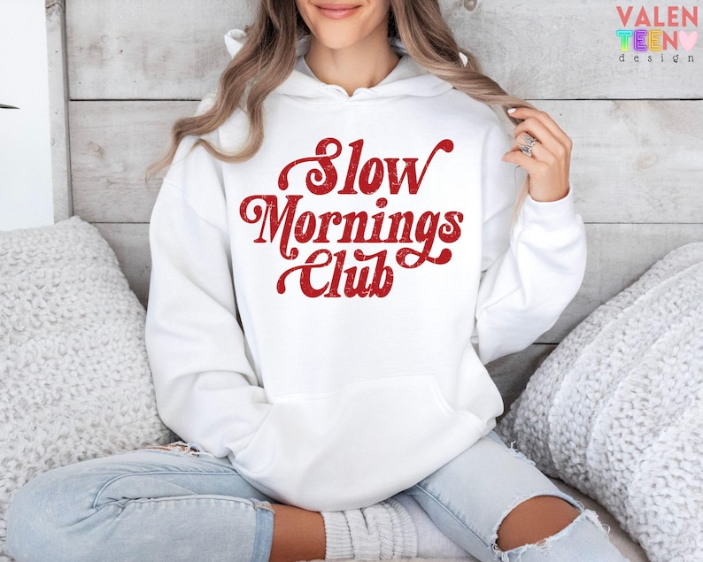 May include: White hoodie with a front pocket and the text "Slow Mornings Club" in red script. The hoodie is made of a soft material and is suitable for casual wear. The design is printed on the front.
