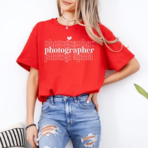 May include: A red t-shirt with the text "photographer" repeated in a stacked pattern with a small heart above the first word. The shirt is being worn by a person with long blonde hair and blue jeans.