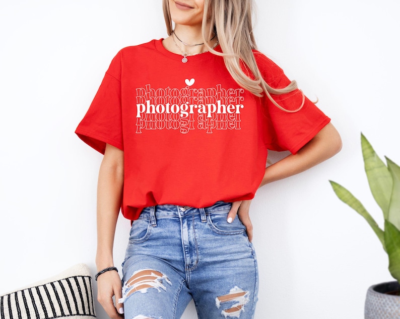 May include: A red t-shirt with the text "photographer" repeated in a stacked pattern with a small heart above the first word. The shirt is being worn by a person with long blonde hair and blue jeans.