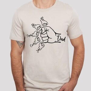 May include: A beige t-shirt with a black graphic of a fist with smaller fists surrounding it. The smaller fists have the names Alex, Sophia, Carlos, and Julia written on them. The larger fist has the word "Dad" written on it.