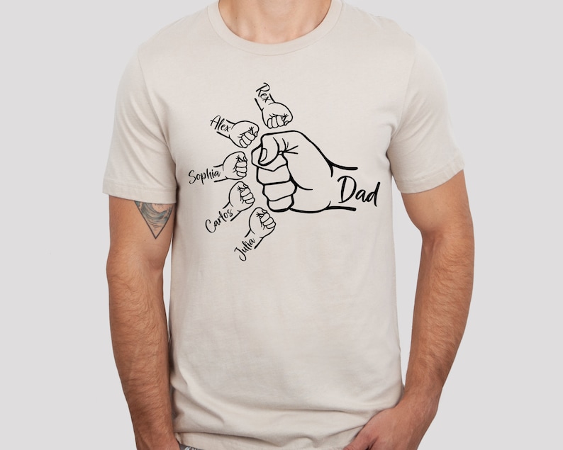 May include: A beige t-shirt with a black graphic of a fist with smaller fists surrounding it. The smaller fists have the names Alex, Sophia, Carlos, and Julia written on them. The larger fist has the word "Dad" written on it.