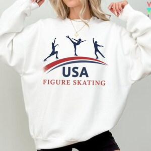 May include: White sweatshirt featuring a graphic of three figure skaters in silhouette, with the text "USA FIGURE SKATING" in navy and red. The design includes a curved red and blue line. The sweatshirt has long sleeves.