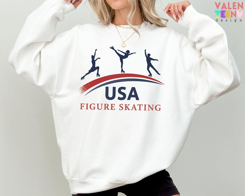 May include: White sweatshirt featuring a graphic of three figure skaters in silhouette, with the text "USA FIGURE SKATING" in navy and red. The design includes a curved red and blue line. The sweatshirt has long sleeves.