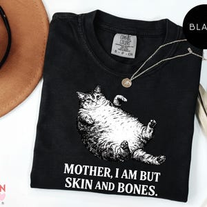 May include: Black t-shirt featuring a graphic of a reclining cat and the text "MOTHER, I AM BUT SKIN AND BONES." A silver necklace with a pendant is on the shirt. A brown hat and decorative plant are in the background.