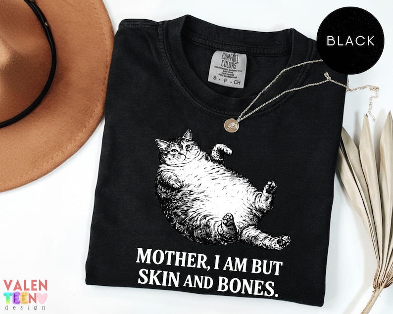 May include: Black t-shirt featuring a graphic of a reclining cat and the text "MOTHER, I AM BUT SKIN AND BONES." A silver necklace with a pendant is on the shirt. A brown hat and decorative plant are in the background.
