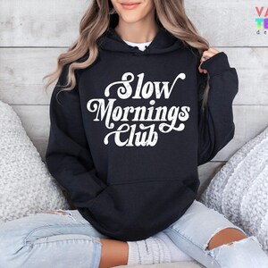 May include: Black hoodie with the text "Slow Mornings Club" in white script. The hoodie has a front pocket and a hood. The person is wearing light blue jeans. The background is a light wood panel.