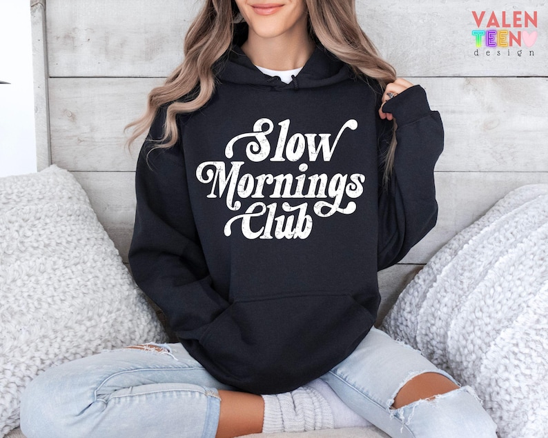May include: Black hoodie with the text "Slow Mornings Club" in white script. The hoodie has a front pocket and a hood. The person is wearing light blue jeans. The background is a light wood panel.