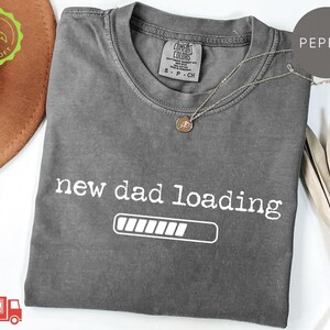 May include: A pepper-colored short-sleeve t-shirt with a crew neckline. The shirt features the white text "new dad loading" above a horizontal progress bar graphic. A small tag inside the collar reads "Comfort Colors" and indicates size small.