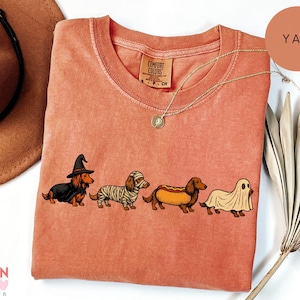 May include: A terracotta-colored short-sleeve t-shirt featuring a graphic of four dachshunds in Halloween costumes: a witch, a mummy, a hot dog, and a ghost. The shirt has a crew neckline and a Comfort Colors brand label visible at the inner collar.