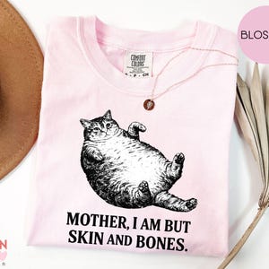 May include: A pale pink t-shirt featuring a black and white drawing of a cat on its back. The text "MOTHER, I AM BUT SKIN AND BONES" is below the cat. A gold necklace is on the shirt.