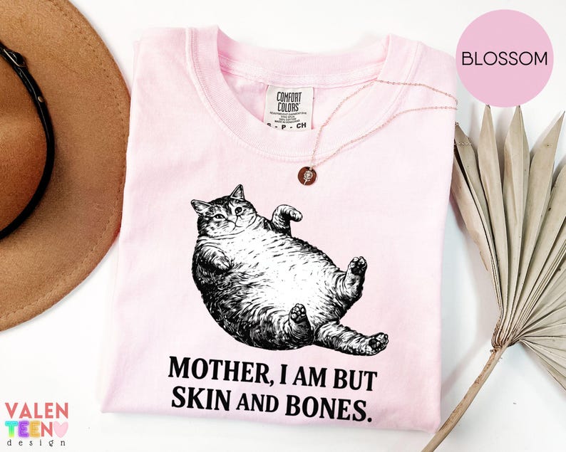 May include: A pale pink t-shirt featuring a black and white drawing of a cat on its back. The text "MOTHER, I AM BUT SKIN AND BONES" is below the cat. A gold necklace is on the shirt.
