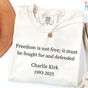 May include: Ivory-colored t-shirt with the quote "Freedom is not free; it must be fought for and defended" attributed to Charlie Kirk (1993-2025). The shirt has a crew neck and short sleeves. A gold necklace with a pendant is draped on the shirt.