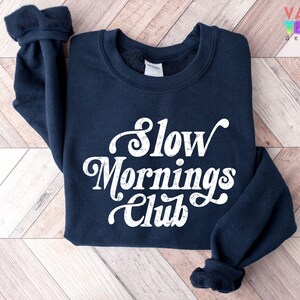 May include: A navy blue sweatshirt with the text "Slow Mornings Club" in white script. The sweatshirt has a crew neck and long sleeves with ribbed cuffs. The Valen Teen Design logo is in the upper right corner.