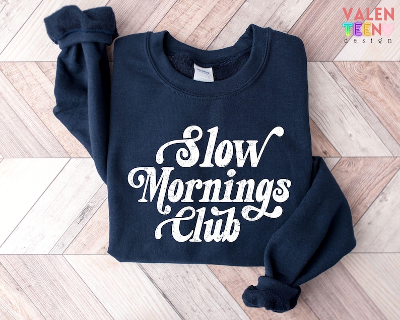 May include: A navy blue sweatshirt with the text "Slow Mornings Club" in white script. The sweatshirt has a crew neck and long sleeves with ribbed cuffs. The Valen Teen Design logo is in the upper right corner.
