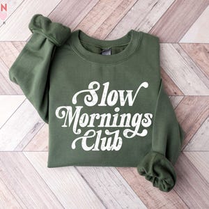 May include: An olive green sweatshirt with the text "Slow Mornings Club" in a white, cursive font. The sweatshirt is folded on a light brown, chevron-patterned wooden surface. The Valen Teen Design logo is in the top left corner.