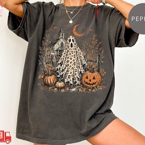 May include: A charcoal gray t-shirt featuring a graphic print of a ghost with a leopard print pattern, standing in front of a church and a wooden fence. The scene includes two orange pumpkins, fallen leaves, and a crescent moon in the night sky.