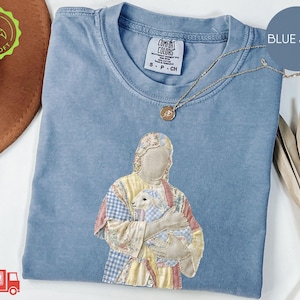 May include: Blue jean colored long-sleeve shirt with an embroidered design of a person holding a lamb. The design features patchwork-style fabric in pastel colors. A gold necklace with a pendant is draped across the shirt.
