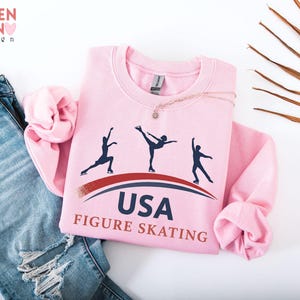 May include: A light pink sweatshirt with the text "USA FIGURE SKATING" and silhouettes of ice skaters. The sweatshirt is folded on top of a pair of blue jeans. The brand "VALENTEEN design" is in the top left corner.