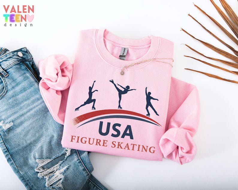 May include: A light pink sweatshirt with the text "USA FIGURE SKATING" and silhouettes of ice skaters. The sweatshirt is folded on top of a pair of blue jeans. The brand "VALENTEEN design" is in the top left corner.