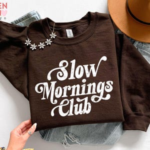 May include: A brown sweatshirt with the text "Slow Mornings Club" in white script. A silver flower necklace is draped on the neckline. A brown hat and blue jeans are in the background.
