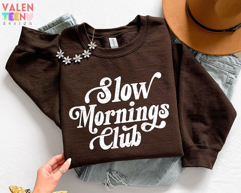 May include: A brown sweatshirt with the text "Slow Mornings Club" in white script. A silver flower necklace is draped on the neckline. A brown hat and blue jeans are in the background.