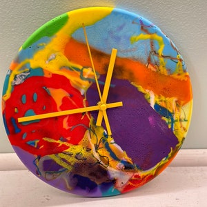 Puede incluir: Un reloj abstracto colorido con manecillas amarillas. La esfera del reloj está hecha de resina y presenta un patrón de remolino de rojo, naranja, amarillo, verde, azul y morado.