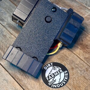 May include: A black and gray electronic device with a round button and a sticker that says "Dirt Farmer Industries".