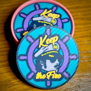 May include: Two round coasters with a blue, purple, and yellow design. The design features a cartoon sailor with a captain's hat and the text "Keep the Fire".