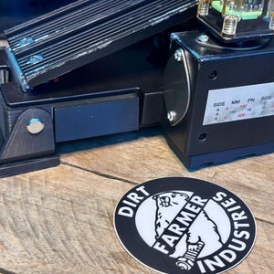 May include: Black foot pedal with a metal plate and a black box with a label that reads "SIDE MM PN SIDE MM PN A 150 18 C 150 18 A 400 18 C 400 18 A 600 18 C 600 18". The pedal is on a wooden surface with a black and white sticker that reads "DIRT FARMER INDUSTRIES".