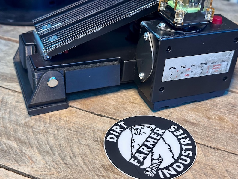 May include: Black foot pedal with a metal plate and a black box with a label that reads "SIDE MM PN SIDE MM PN A 150 18 C 150 18 A 400 18 C 400 18 A 600 18 C 600 18". The pedal is on a wooden surface with a black and white sticker that reads "DIRT FARMER INDUSTRIES".